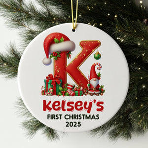 A white ornament with a red letter "K" and a gnome, along with various Christmas-themed elements such as presents and a ball. The ornament is hanging on a Christmas tree, and it is labeled as "Kelsey's First Christmas 2025".