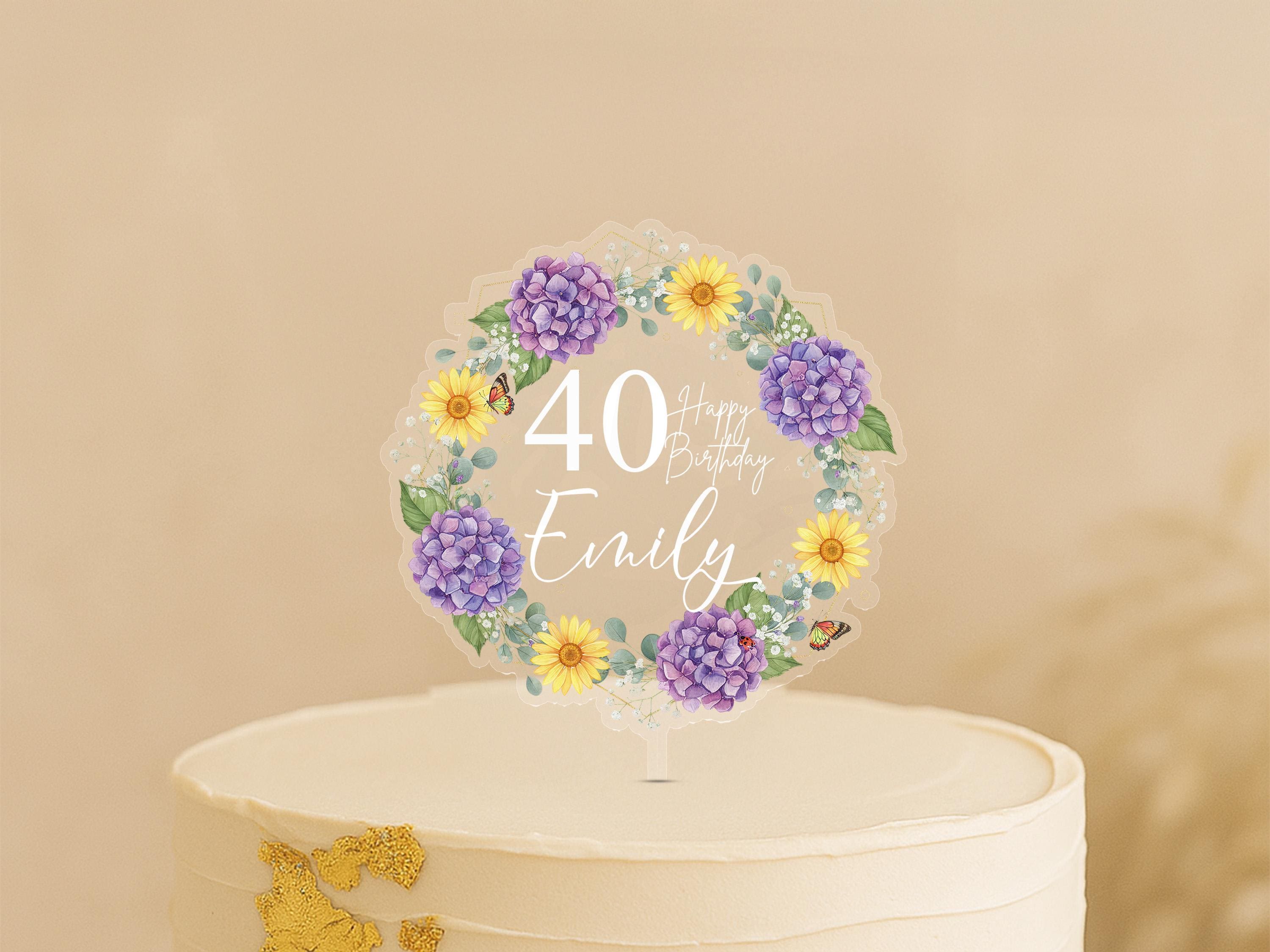 A white cake with a wreath-shaped birthday topper featuring purple and yellow flowers, along with the text "Happy Birthday Emily".