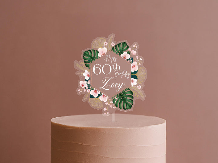 A cake topper with the text "Happy 60th Birthday Lucy" surrounded by a floral design.