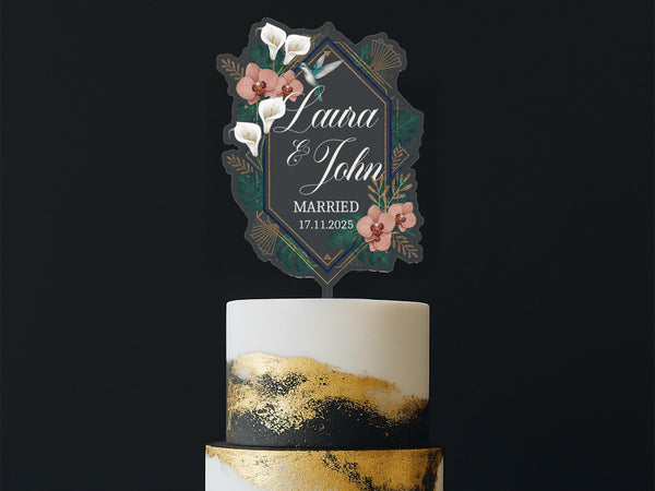 Personalised Art Deco wedding cake topper featuring geometric gold and emerald green frame with white calla lilies, terracotta orchids, tropical palm leaves and hummingbird, custom names Laura and John, Married 17.11.2025, displayed on elegant gold and black painted cake