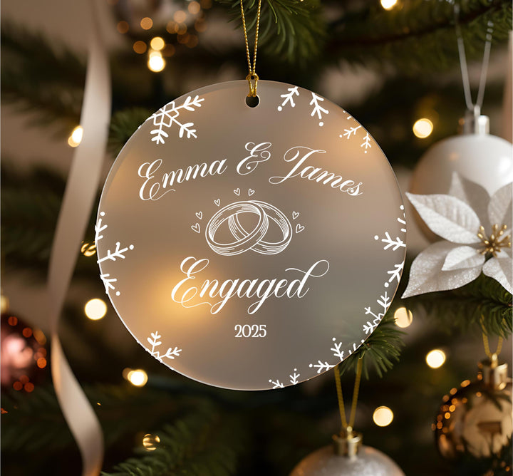 Personalised engagement Christmas ornament with couple names Emma and James, wedding rings, hearts and snowflake border on frosted acrylic, hanging on Christmas tree with tartan ribbon and gold baubles