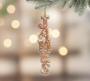A decorative ornament made of gingerbread letters spelling out the name "JAMES" hanging from a branch of a pine tree.