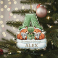 A Christmas ornament featuring two cartoon foxes wearing Santa hats and scarves, with a green letter "A" in the center. The ornament is hanging on a decorated Christmas tree.