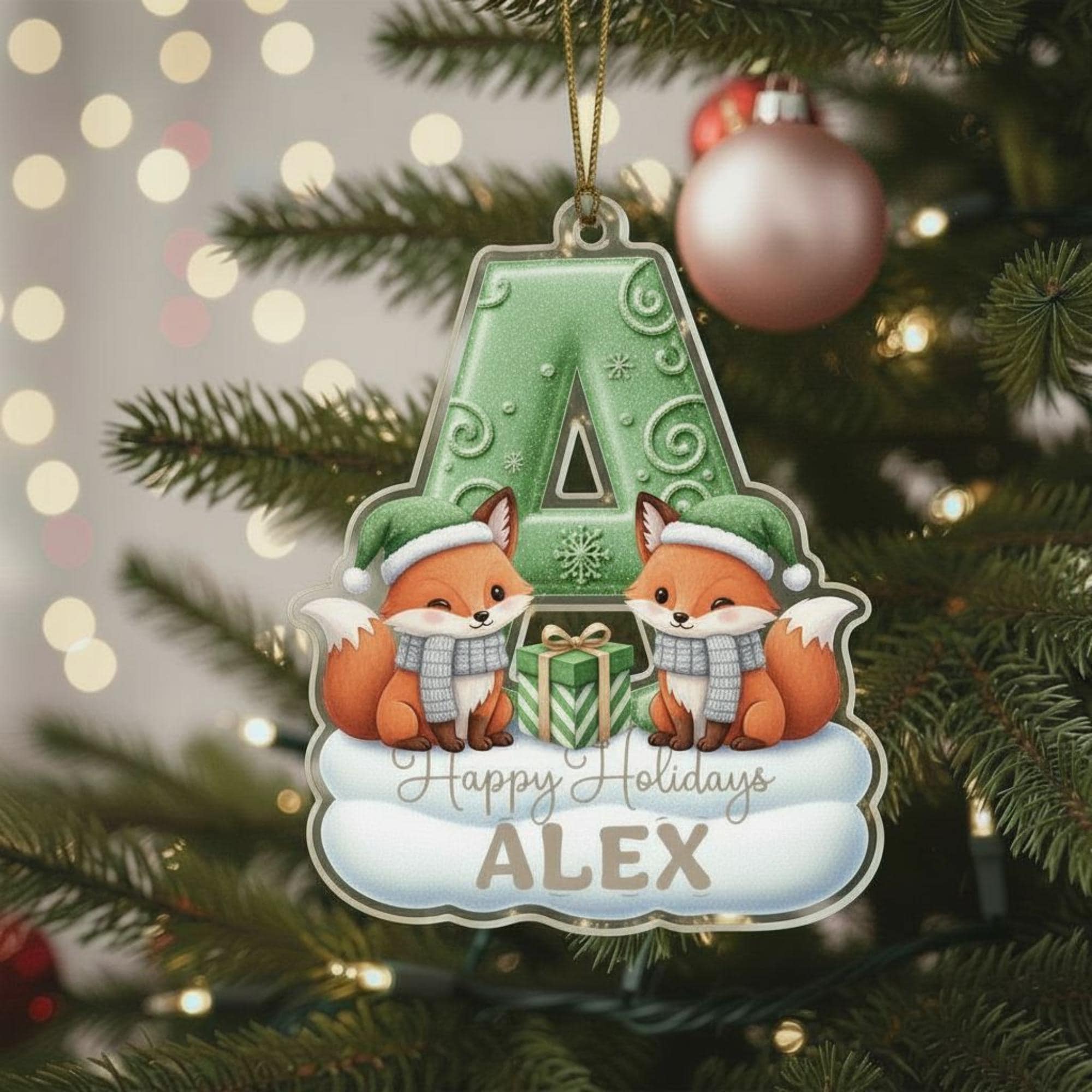 A Christmas ornament featuring two cartoon foxes wearing Santa hats and scarves, with a green letter "A" in the center. The ornament is hanging on a decorated Christmas tree.