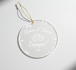 A clear glass ornament with a gold string attached, featuring the names 'Emma & James' and the year '2025' along with a snowflake design.
