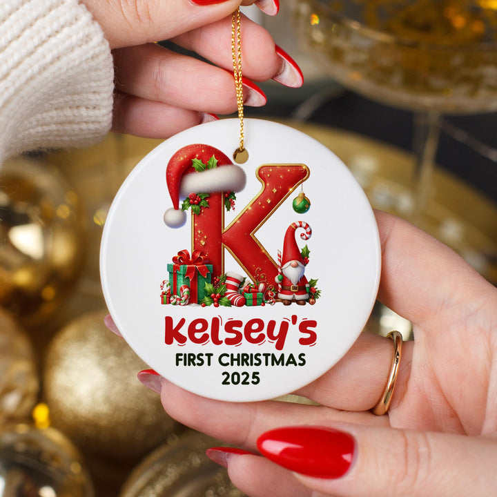 Personalised baby first Christmas ornament featuring letter K with Santa hat, Christmas gnome, presents, candy canes and custom name Kelsey&#39;s First Christmas 2025 held in hands