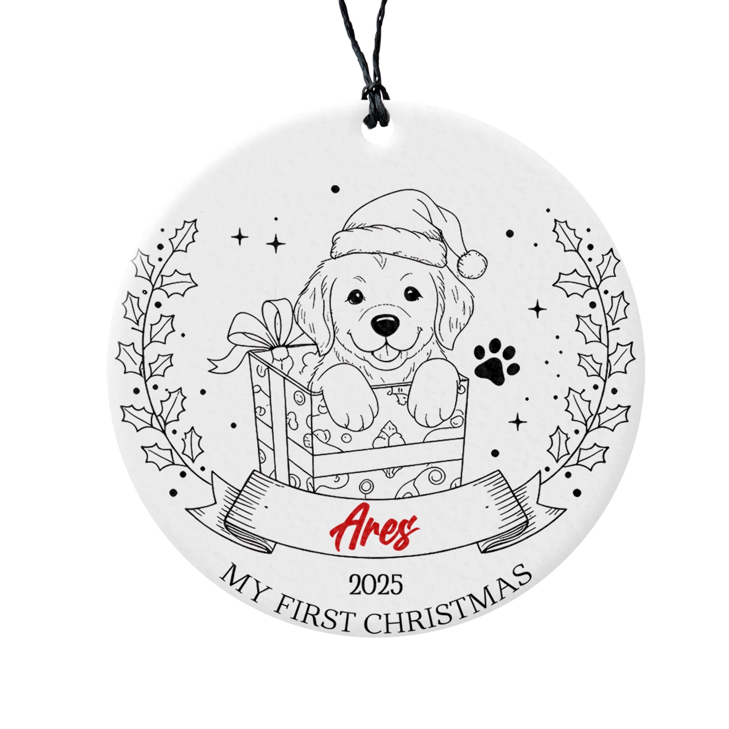 A white ornament with a black and white illustration of a dog wearing a Santa hat, sitting inside a gift box, and surrounded by holly leaves and stars. The ornament has a banner that says "Ares" and "My first Christmas" below the illustration.