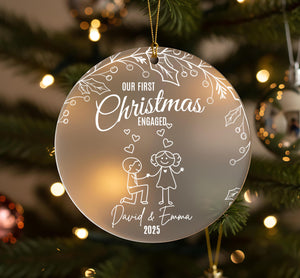 A Christmas ornament with a couple of people, one kneeling and the other standing, surrounded by hearts and leaves. The ornament has a message that reads "Our first Christmas engaged David & Emma" and the year "2025".