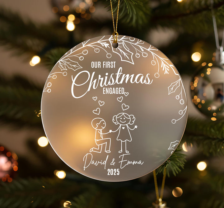 A Christmas ornament with a couple of people, one kneeling and the other standing, surrounded by hearts and leaves. The ornament has a message that reads &quot;Our first Christmas engaged David & Emma&quot; and the year &quot;2025&quot;.