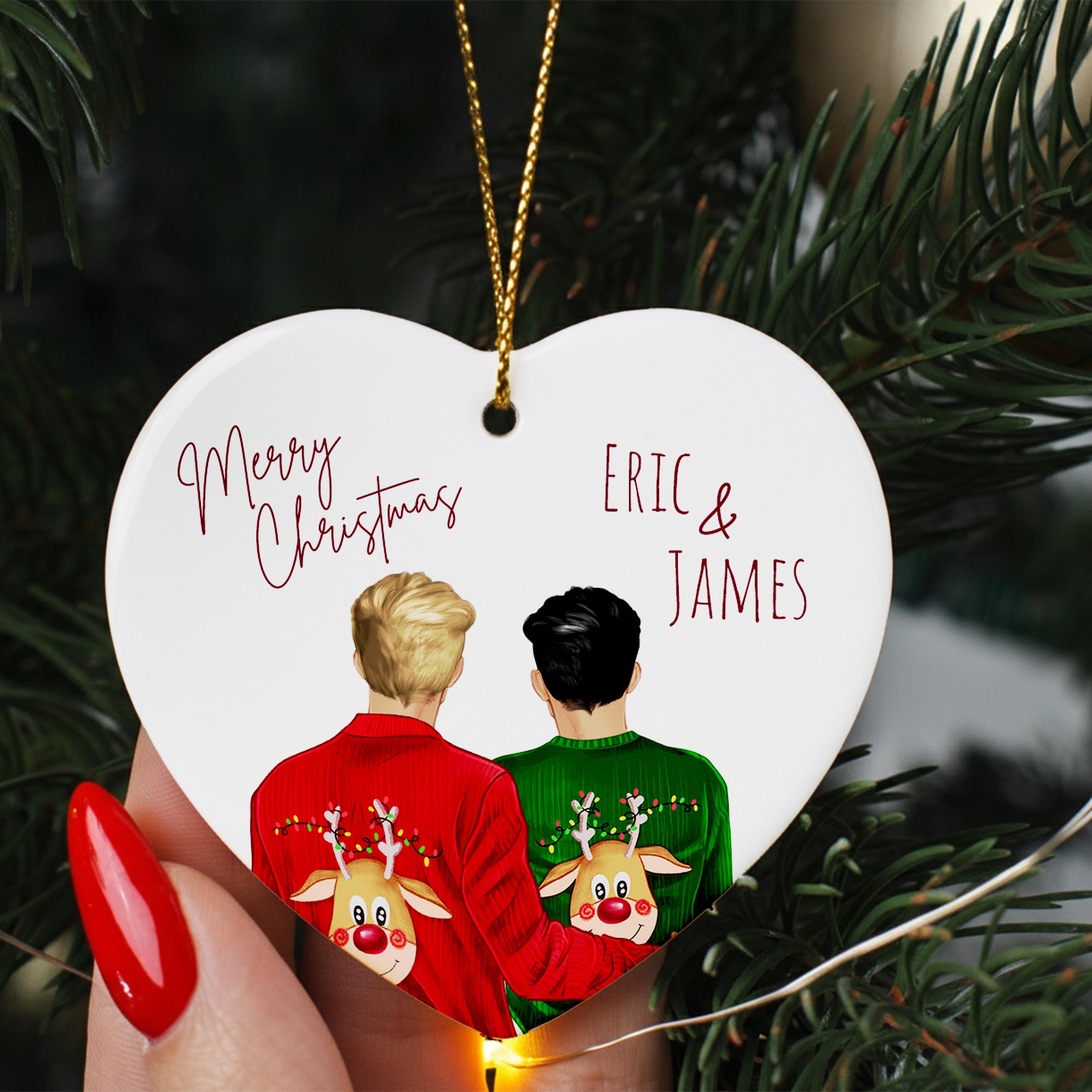 A heart-shaped ornament with two people wearing Christmas sweaters, one in red and the other in green, with a deer face on their backs. The ornament is hanging from a string, and there is a "Merry Christmas" message and the names "ERIC & JAMES" written on it.