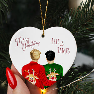 A heart-shaped ornament with two people wearing Christmas sweaters, one in red and the other in green, with a deer face on their backs. The ornament is hanging from a string, and there is a "Merry Christmas" message and the names "ERIC & JAMES" written on it.