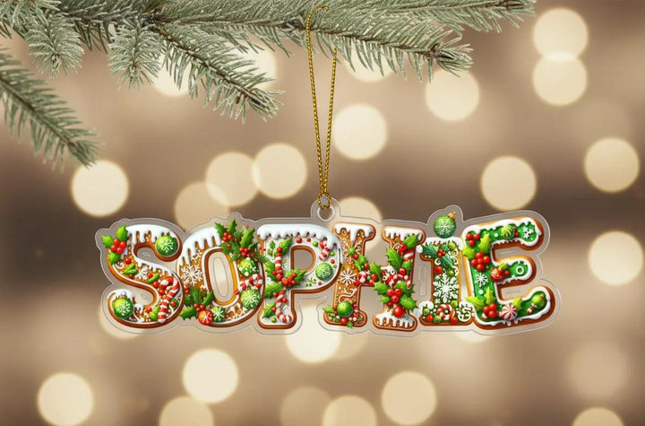 A clear ornament shaped like the name &quot;Sophie&quot; made of gingerbread cookies, berries, and other festive decorations, hanging on a branch with a blurred background of lights.