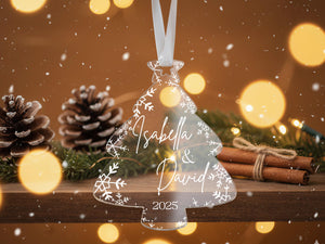 A clear ornament shaped like a Christmas tree with the names "Isabella & David" and the year "2025" written on it, hanging from a ribbon. The ornament is surrounded by pine cones, cinnamon sticks, and other holiday decorations, with a blurred background of lights.