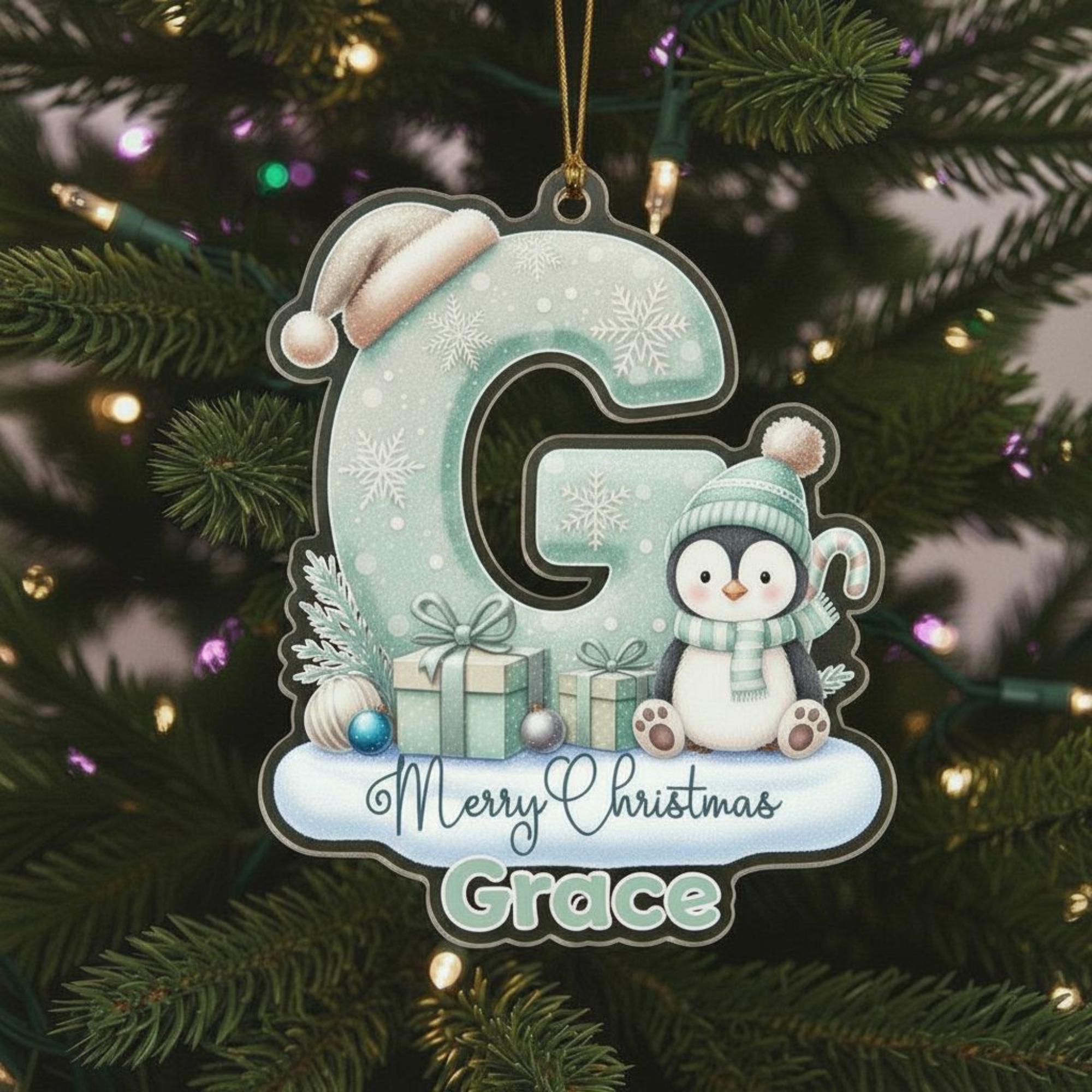 A Christmas ornament shaped like the letter "G" with a penguin and presents, hanging on a tree with lights.