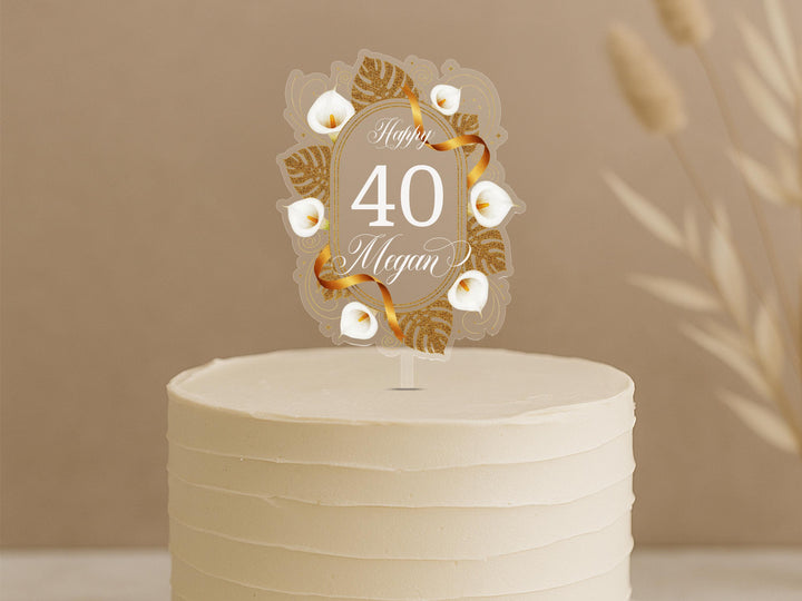 A white cake with a decorative gold and white floral topper that says &quot;Happy 40 Megan&quot; on it.