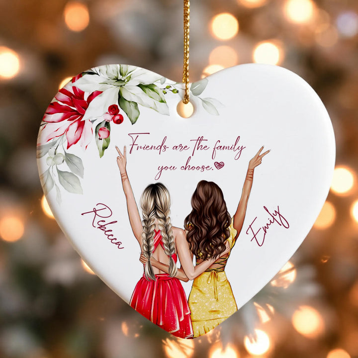 A heart-shaped ornament with two women in colorful dresses, one with blonde hair and the other with brown hair, standing together and smiling. The ornament has a floral design and the text &quot;Friends are the family you choose&quot; written on it.