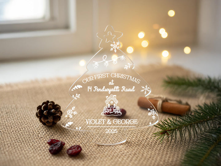 A clear glass Christmas tree ornament with the text &quot;Our first Christmas at 11 Pendwyatt Road&quot; and &quot; Violet & George 2005&quot; on it, surrounded by pine branches, cranberries, and a star decoration.