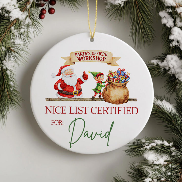 A white ornament with a Santa Claus and an elf, along with a bag of presents, hangs on a Christmas tree.