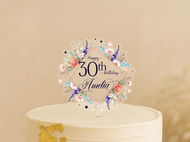 A white cake with a decorative floral topper that says "Happy 30th Birthday Amanda" on it.