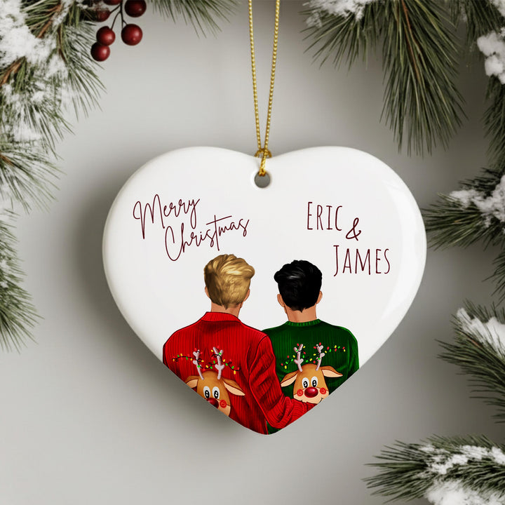A heart-shaped ornament with two people wearing Christmas sweaters and reindeer antlers, surrounded by pine branches and red berries.