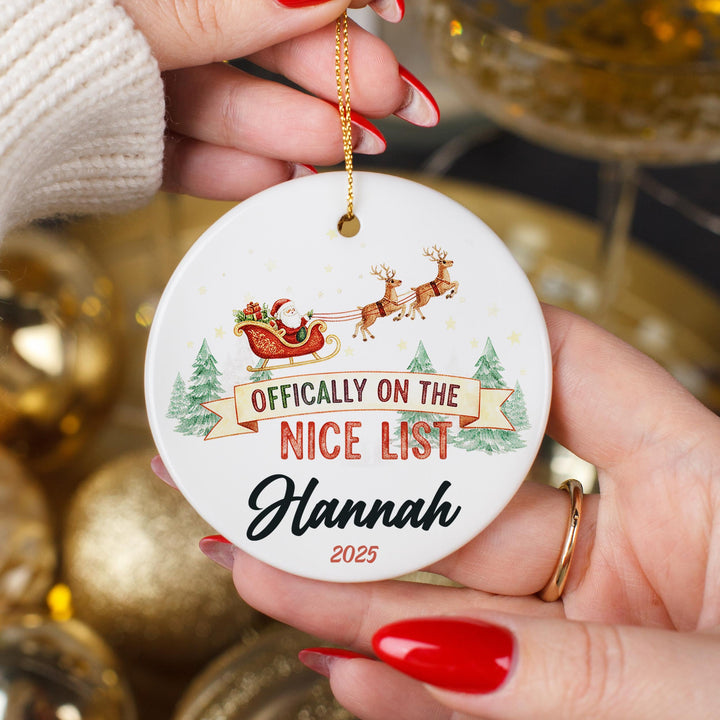 Personalised ceramic Christmas ornament featuring Santa&#39;s sleigh with reindeer flying over pine trees, text Officially on the Nice List with custom name Hannah 2025, held in hands showing glossy finish