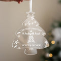 Personalised clear acrylic Christmas tree ornament engraved with Our First Christmas at 4 Pendwyallt Road, names Violet & George 2025, featuring snowflakes and house design, hanging on Christmas tree with lights