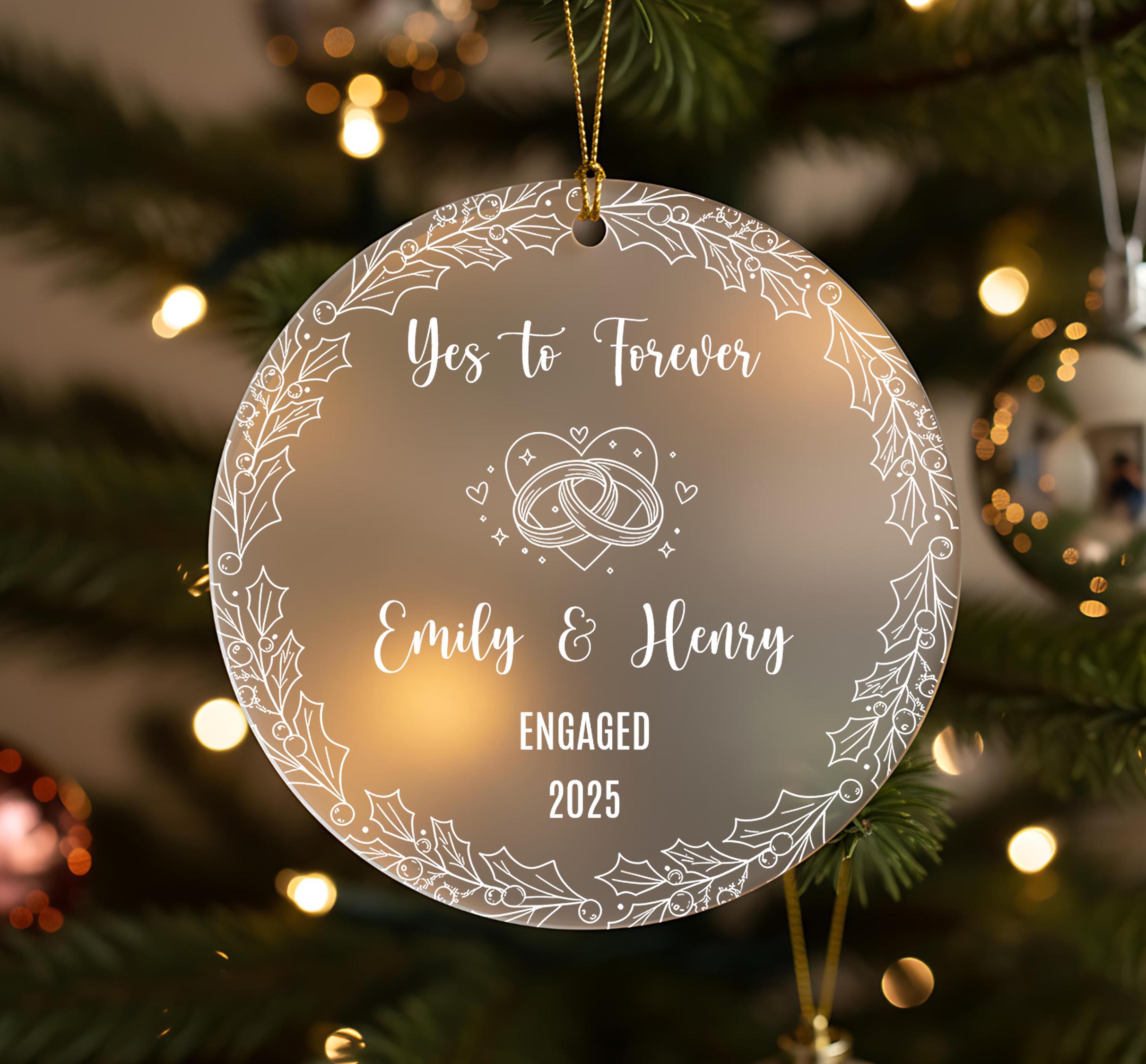 A clear ornament with a white floral design hangs on a Christmas tree, displaying the text "Yes to Forever Emily & Henry Engaged 2020".