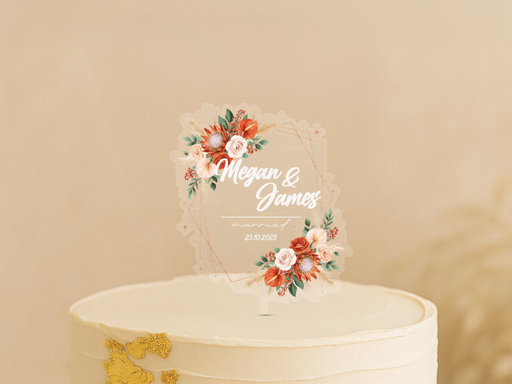 A white wedding cake with a clear floral topper displaying the names &quot;Megan & James&quot; and the date &quot;23/03/2023&quot;.