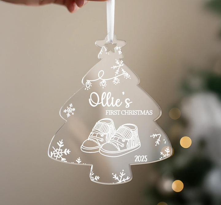 A hand holding a clear ornament shaped like a Christmas tree with a personalized message and a pair of baby shoes.