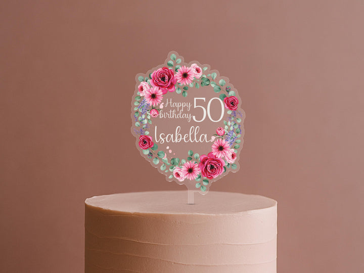 A birthday cake with a clear wreath-shaped topper featuring pink flowers and the text &quot;Happy Birthday Isabella&quot; on it.