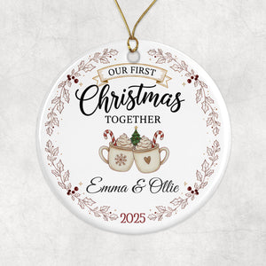 A white ornament with a festive design, featuring a couple enjoying hot chocolate together. The text on the ornament reads "Our first Christmas together" and "Emma & Ollie" along with the year "2025".