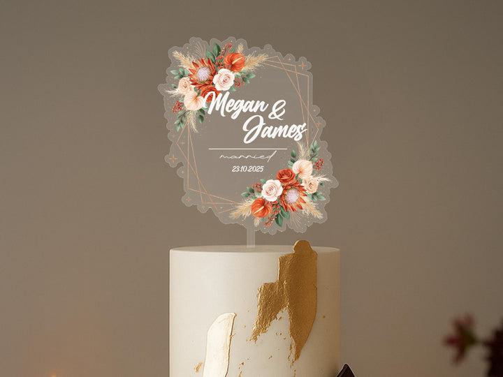 A white cake with a decorative floral design on top, featuring the names &quot;Megan & James&quot; and the date &quot;23.10.2025&quot; on it.
