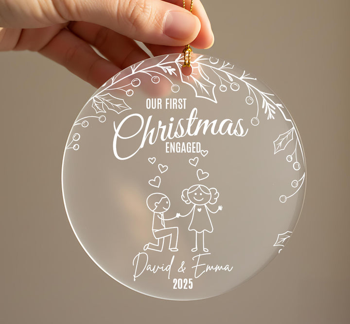 Personalised first Christmas engaged ornament held in hand showing frosted acrylic design with couple proposal illustration, holly leaves, berries and hearts, engraved with names David and Emma 2025