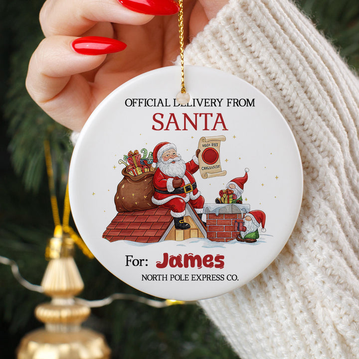 A person&#39;s hand holding a white ornament with a Santa Claus illustration and text.