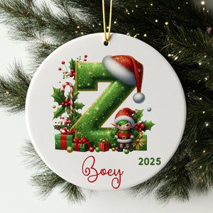 A white ornament with a green glittery letter "Z" and a small elf wearing a Santa hat, surrounded by Christmas decorations such as presents and berries.