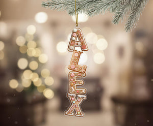 A gingerbread house ornament with the name "ALEX" made of gingerbread pieces is hanging from a branch.