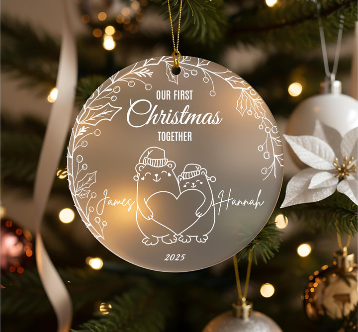 A Christmas ornament with a couple of bears holding hands, surrounded by a wreath and other ornaments, with the text &quot;OUR FIRST CHRISTMAS TOGETHER&quot; and &quot;James Hannah&quot; written on it.
