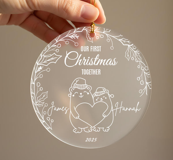 Personalised first Christmas together frosted acrylic ornament held in hand showing two cute bears wearing winter hats holding a heart, engraved text Our First Christmas Together, couple names James and Hannah on either side, 2025, surrounded by holly leaf and berry border with gold ribbon