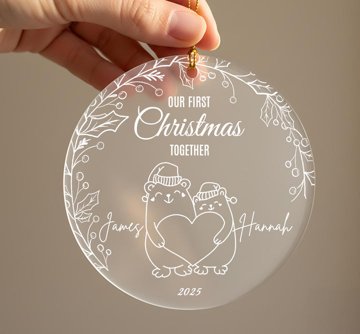 Personalised first Christmas together frosted acrylic ornament held in hand showing two cute bears wearing winter hats holding a heart, engraved text Our First Christmas Together, couple names James and Hannah on either side, 2025, surrounded by holly leaf and berry border with gold ribbon