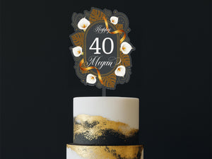 A white cake with gold and black designs, and a decorative topper that says "Happy 40 Megan" in a gold and black design.