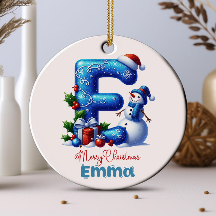 A white ornament with a snowman and Christmas decorations, including a gift box and holly leaves, is hanging from a string.