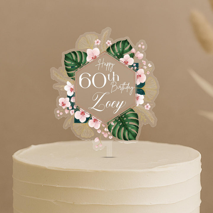 A white cake with a clear hexagonal topper featuring pink flowers and green leaves, along with the text "Happy 60th Birthday Zoey".