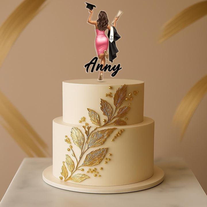 A two-tiered white cake with gold leaf decorations and a graduation cap topper featuring a woman in a pink dress.