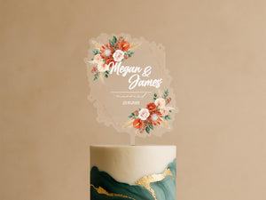 A wedding cake topper with the names 'Megan & James' and the date '23.10.2021' displayed on it, surrounded by a floral design.