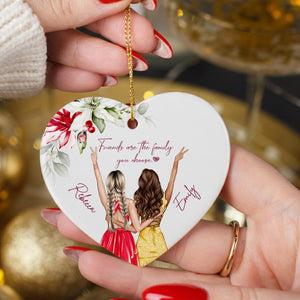 A heart-shaped ornament with two women in colorful dresses, one with blonde hair and the other with brown hair, both with their arms raised in a celebratory manner. The ornament is being held by a person's hand, and there is a wine glass visible in the background.