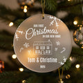 Personalised first Christmas in new home frosted acrylic ornament hanging on Christmas tree with gold ribbon showing engraved text Our First Christmas in Our New Home, house illustration with presents and candy canes, couple names Tom and Christine 2025, decorated with snowflakes and winter sprigs against bokeh lights