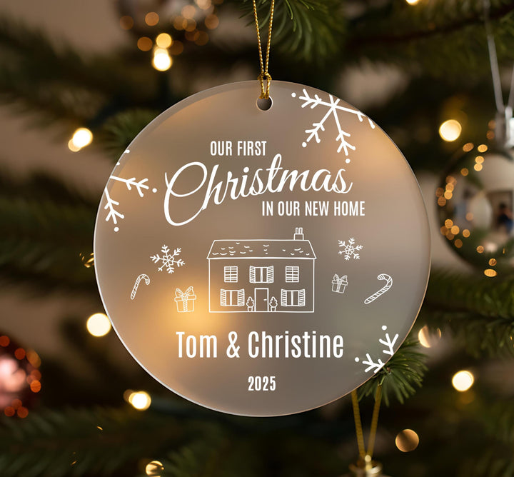 Personalised first Christmas in new home frosted acrylic ornament hanging on Christmas tree with gold ribbon showing engraved text Our First Christmas in Our New Home, house illustration with presents and candy canes, couple names Tom and Christine 2025, decorated with snowflakes and winter sprigs against bokeh lights