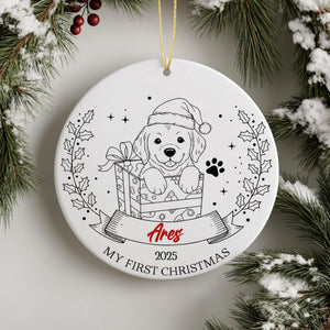 A white ceramic ornament with a black and white illustration of a dog wearing a Santa hat, sitting in a gift box, surrounded by a wreath and berries. The ornament has a ribbon that says "Ares" and "MY FIRST CHRISTMAS".
