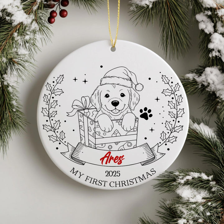 A white ceramic ornament with a black and white illustration of a dog wearing a Santa hat, sitting in a gift box, surrounded by a wreath and berries. The ornament has a ribbon that says &quot;Ares&quot; and &quot;MY FIRST CHRISTMAS&quot;.