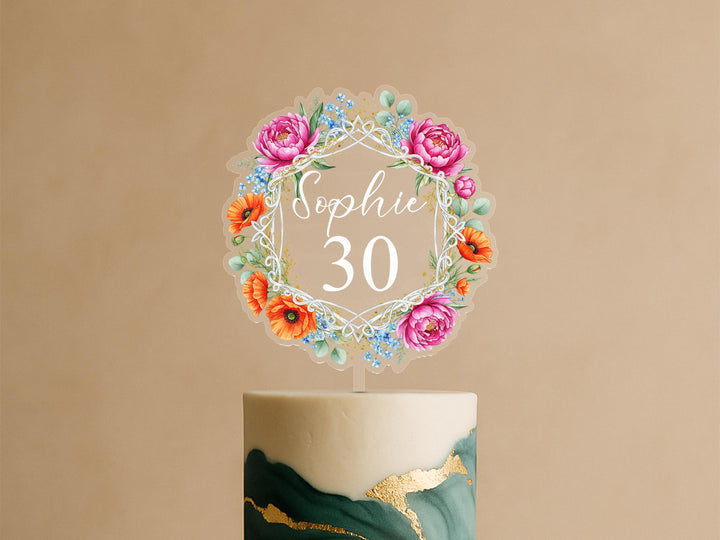 Personalised celebration cake topper featuring geometric frame with pink peonies, orange poppies, blue wildflowers and custom name Sophie 30 on elegant green and gold celebration cake